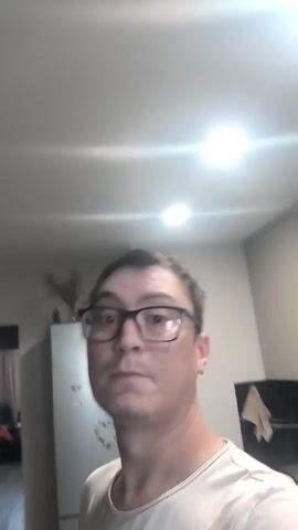 A medium shot shows a man wearing glasses and a light-colored t-shirt in a kitchen. He is looking directly at the camera with a neutral expression. The background includes a refrigerator with magnets on it and kitchen cabinets. A light fixture is visible on the ceiling, casting bright light. The video appears to be a selfie taken by the man.