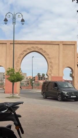 The video shows a cityscape featuring a large, arched, beige structure in the foreground. A scooter is parked on the left side of the frame. A motorcycle with a delivery driver and a yellow delivery box passes through the archway. A black van is parked on the right. Through the archway, there's a view of a city with buildings and palm trees, possibly under construction. The sky is partly cloudy. There's a lamppost to the left of the archway, and some graffiti on the side of the arch. The street is paved with rectangular stones.