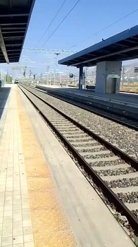 A train station platform with two sets of train tracks. On the left, a person in a dark blue uniform stands on the platform. In the distance, a train is approaching on the tracks. On the right, a modern train station canopy with an elevator is visible. The sky is clear and blue. A person on the left appears to be talking to someone out of frame. The train is in motion.