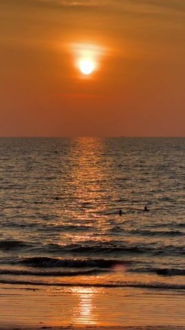 The video shows a sunset over the ocean, with the sun appearing as a bright white circle in an orange sky. The water reflects the sunlight, creating a shimmering path towards the shore. Small waves lap at the beach in the foreground. There are three dark shapes in the water, possibly people swimming or floating.