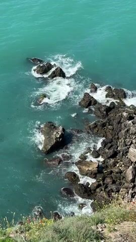 The video shows a high-angle view of turquoise water crashing against a rocky shoreline. The rocks are dark and jagged, forming small inlets and outcrops. White foam from the waves contrasts with the blue-green water. Patches of low-lying vegetation, including some small red flowers, are visible along the cliff edge. The water appears slightly choppy, and small bits of debris float on the surface.