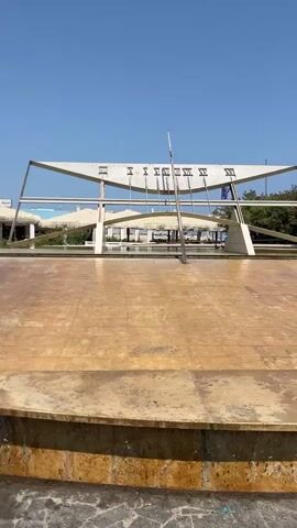 A horizontal video captures a sundial in an outdoor setting under a clear blue sky. The sundial is a prominent feature, with its gnomon (the part that casts the shadow) angled upwards and the hour markers displayed in Roman numerals. The sundial is mounted on a modern, angular metal structure. In the foreground, there is a circular, slightly sloped, weathered concrete or stone area, possibly part of a fountain or plaza. Water is seen trickling down the side of this circular platform, suggesting a water feature. In the background, there are some buildings with awnings and trees. The overall impression is a sunny day at a public space.