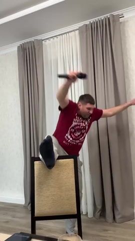 A man in a maroon t-shirt and grey sweatpants dances energetically in what appears to be a dining room. He steps on a chair with one foot, holding a black object like a remote control in his hand, then loses his balance and comes down laughing. The room is decorated with beige and white curtains, and patterned wallpaper. Parts of a dining table and other people are visible in the foreground. Upbeat music plays in the background.