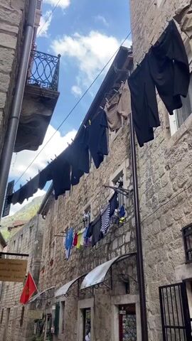 This video captures a narrow, ancient cobblestone alleyway in Kotor, Montenegro, during the daytime. The alley is bordered by tall, historic stone buildings on both sides.

A prominent feature is the laundry hanging on clotheslines stretched across the alley between the buildings. Dark pants are seen hanging on a higher line, while a lower line displays a variety of colorful clothes, including a striped shirt, blue, yellow, and red garments, some dangling from a window on the right-hand building.

In the foreground, a young boy stands in an open doorway on the right side of the alley, looking out. Further down the alley, several other people are visible, some walking and others gathered, suggesting a lively pedestrian area.

Notable details include a metal balcony on a building to the left, a drainage pipe running down the side of the left building, and a red flag, resembling the Montenegrin flag, hanging from a building on the right. A wooden sign on the left building clearly reads: "Roman Handmade Sandals & Shoes Man & Women Fashion". Another sign with "2.00" is visible near the boy's doorway. The sky above is partly cloudy. The overall scene depicts a charming, old-world street with everyday life unfolding.