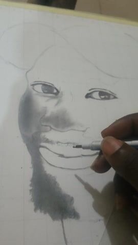 A hand is shown holding a pen and drawing a portrait of a person with curly hair. The drawing is in black and white and is on a white piece of paper. The hand is darker-skinned. The pen is silver with black and blue accents.  The portrait is unfinished, but the eyes, nose, and part of the hair are visible. The background is blurry.