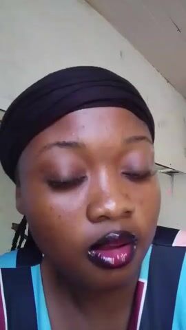 A medium shot captures a woman looking at the camera. She has medium brown skin, dark eyes, and a smooth complexion. She has dark false eyelashes and a subtle eye shadow. Her eyebrows are defined, and she has a dark reddish-brown lipstick. She has a black headwrap on her head. Her striped shirt has colors of blue, purple, and black. A person is visible in the background, out of focus.