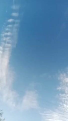 The video shows a sunrise behind a distant hilltop silhouette. In the foreground, a telephone pole with lines stretches into the sky, which is filled with textured, rippled cloud formations. The sky transitions from a clear blue above to a muted, brighter hue near the horizon where the sun is rising. The overall shot is taken from a low angle, emphasizing the height of the pole and the expanse of the sky.  The silhouette of a person is visible standing on the hilltop.