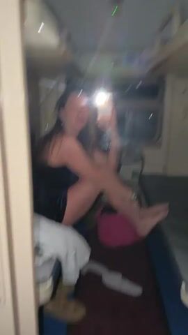 A woman is seen taking a selfie in a mirror inside a train compartment. She is sitting cross-legged on the lower bunk with her phone in her right hand, facing the mirror. She is wearing a black dress. A pink bag is next to her. There are two bunks in the compartment, one above her and the other across from her. The window is behind her. There is a pair of sandals on the floor. The lighting is dim, mainly from the phone's flash. The train compartment appears small and functional. The image appears to be filmed on a cell phone, held in front of the mirror.