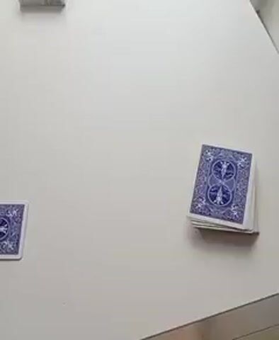 This is a close-up shot of a white table with several decks of playing cards. In the upper left corner, a deck of cards is visible. To the left of the frame, a single playing card is lying face up. In the lower right portion of the frame, a deck of playing cards is standing upright, with the backs of the cards facing the camera. The cards have a blue and white pattern. The lighting is bright, and the focus is sharp.