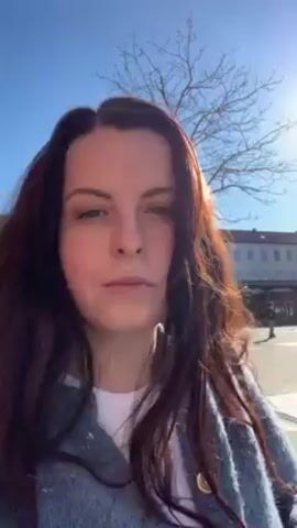 A woman with long brown hair walks outside on a sunny day. She is wearing a light blue cardigan over a white top. The sun is directly behind her, creating a glare. There are buildings, trees, and lampposts in the background.