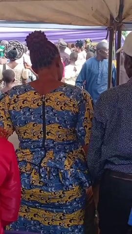 The video depicts a gathering of people, likely at an event or celebration.  The setting appears to be outdoors under a tent-like structure.  Many people are standing and watching a performance. Performers are wearing matching outfits and appearing to dance.  There are tables with bottles and other objects on them. People in the crowd are wearing a variety of casual and dressier clothing.  Some are taking photos or recording video.