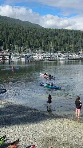 This is a vertical, medium shot video of a harbor with several people kayaking in the water and a few people standing on the shore. In the foreground, two young girls are standing in shallow water on a pebbly beach. Behind them, three kayaks are moving across the water. The first kayak, a blue and green one, has a person in a blue shirt and a red helmet paddling. The second kayak, a white one, has two people paddling, one in a red shirt and one in a blue shirt. The third kayak, a blue one, has a person in a red shirt paddling. In the background, a marina is filled with sailboats. A tree-covered mountain rises behind the marina under a partly cloudy sky. The lighting suggests it is daytime.