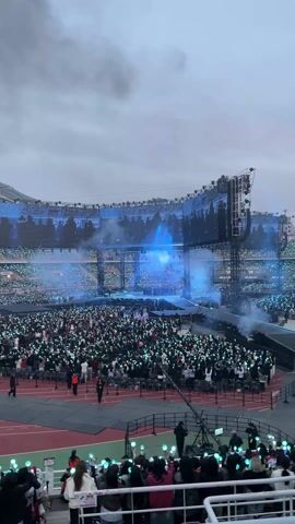 The video shows a large crowd of people at a stadium concert. They are holding light sticks, and there is a stage with large screens. The screens show images of the performers. The sky is overcast and grey. The stadium is filled with people, and there are cameras and other equipment set up on the sidelines.