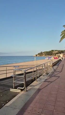 The video shows a paved walkway next to a sandy beach. A metal railing is visible along the walkway, which appears to slope upwards. The sea is calm and blue, meeting the sandy beach. In the background, a hill covered with trees and some buildings can be seen. There are a few beach chairs on the sand. The sky is clear and blue. The video is shot in landscape orientation.