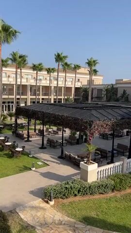 The video pans across a resort courtyard with palm trees and lush greenery. A covered seating area with tables and chairs is visible, as well as a white balustrade along the edge of the courtyard. In the background, a multi-story hotel building is situated. The sky is clear and blue, indicating a sunny day.