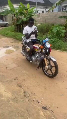 The video shows a person sitting on a motorcycle and looking at their phone. The motorcycle is parked on a dirt road, which has several puddles of water. In the background, there are some buildings, trees, and a car. Later in the video, a man is seen carrying a child and walking down the dirt road. There is also a small white dog sitting on the side of the road.