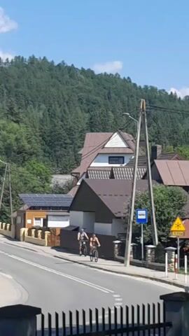 Two individuals on bicycles are riding down a paved road in front of a residential area. Several houses with brown roofs are visible, some with solar panels on their roofs. The road has white lane markings. A blue car is driving on the road, and a person on a motorcycle is also present. There are traffic signs, including a bus stop sign and a yellow warning sign. A densely wooded hill is in the background under a clear blue sky.