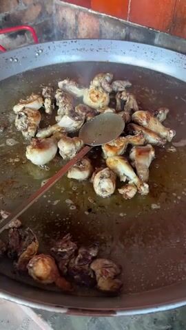 The video shows a large paella pan filled with pieces of chicken being cooked in oil. A large slotted spoon is used to stir the chicken. Someone pours a brown liquid from a glass bottle into the paella. The camera moves to an outdoor patio area, where a man in white shorts and a white shirt walks across the patio carrying a plate. A dog is lying on the tile patio. Other people are sitting at a table in the background. Pink flowers are visible in planters along the patio.