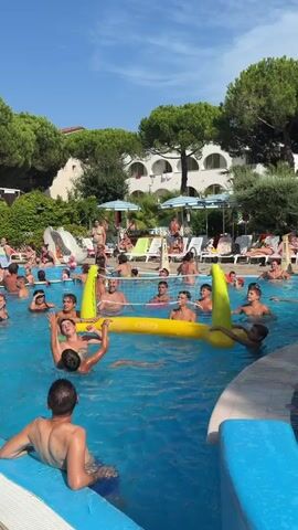 This is a daytime video taken from above the surface of a bright blue swimming pool. A group of people, primarily adults and children, are playing volleyball in the pool. An inflatable yellow volleyball net is set up in the middle of the pool. The players are actively engaged in hitting the ball over the net. Some people are standing in the water, while others are partially submerged. In the background, there are lounge chairs with people relaxing, umbrellas, lush green trees, and a building with white walls and arched windows. The sky is clear and blue. The overall atmosphere appears to be lively and recreational.