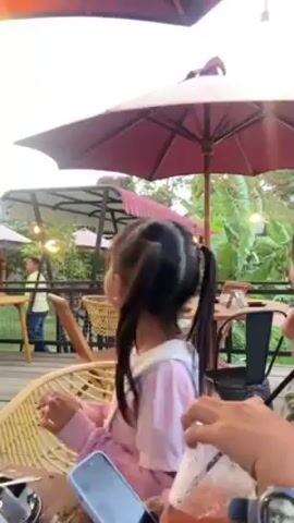 The video shows two young girls sitting at a table on an outdoor patio. One girl has pigtails and is wearing a pink shirt, while the other is wearing a green top and has a white bow in her hair. The girls appear to be talking and interacting with each other. In the background, there is a view of a mountain range, green fields, and several tables and chairs on the patio. String lights are hanging above the patio area. The weather appears to be overcast. The camera pans from the girls to the scenery and back again.