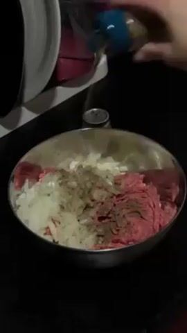 A person is adding spices to a bowl of raw ground beef and chopped onions. The bowl is stainless steel and sits on a black countertop. There are multiple spice containers on the counter near the bowl. A white kitchen appliance is visible on the left.