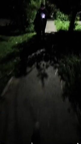 The video shows a first-person perspective of someone riding a bicycle at night on a paved path. The path is lined with grass and trees. The cyclist is moving forward and the camera is facing down the path. A person is briefly visible in the distance, holding a phone that is illuminating their face. The scene is dimly lit by artificial lights from unseen sources and the headlights of the bicycle. The overall impression is of a quiet, nighttime ride through a park or suburban area.