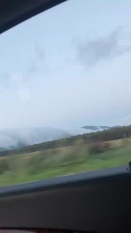 The video shows a view from inside a vehicle. In the first few seconds, the camera captures a landscape with rolling hills, a line of trees, and a band of clouds near the horizon under a pale sky. The scene appears to be a natural landscape. Then, the view shifts to show a roadside commercial establishment. It's a red building with bright signage, possibly a convenience store or gas station, with illuminated blue and red lettering. A white car is parked to the left of the building, and another dark-colored vehicle is visible in the background. The environment appears to be a commercial area situated near a natural landscape, likely observed during a road trip. The lighting suggests it is either early morning or late evening.