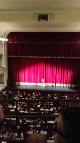The image shows a performance taking place on a stage in a theater. A single person, dressed in what appears to be a costume, stands center stage, illuminated by a spotlight. The stage is framed by a large, red velvet curtain with gold trim. The audience, seated in rows, is visible in the right side of the frame. The theater's ornate architecture is visible in the upper left corner. The lighting suggests the performance is happening indoors, likely in the evening. The overall atmosphere appears to be one of anticipation and engagement from the audience. The person on stage seems to be the focus of attention, possibly mid-performance or about to begin. There is no visible text in the image.