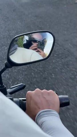 A first-person perspective video shows a right hand gripping a motorcycle handlebar. The primary focus is on the right rearview mirror, reflecting the rider. The rider wears a dark helmet, sunglasses, and a light-colored shirt, and is holding a dark smartphone to film their reflection. The mirror also reflects trees and a blue sky. Outside the mirror, an asphalt road and a grassy verge are visible, indicating movement.
