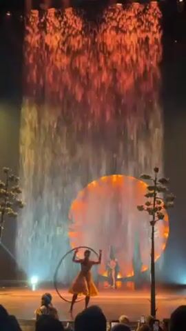 The video shows a performance on a stage with a water curtain effect in the background. There is a large, illuminated orange circle in the middle of the stage, flanked by two tree-like structures. In the foreground, a person in a yellow dress spins a hula hoop. Then, an aerialist descends from the top of the stage on a triangular swing. The audience is visible in the foreground, with some people holding up their phones to record the performance. The lighting creates an overall warm, theatrical atmosphere.