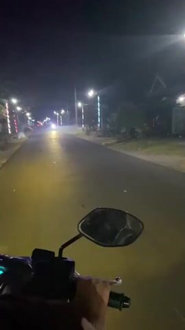 A first-person perspective video shows a person riding a motorcycle at night on a paved road. Streetlights illuminate the road, and decorative lights are visible on poles lining the street. Trees and buildings can be seen on either side of the road. The motorcycle's headlight casts a yellow glow on the road ahead. Another motorcycle with its headlights on passes by in the distance. The video is smooth, indicating a steady ride.