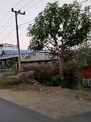 The video shows a view from a moving vehicle on a street. On the right side of the street, there are several houses and buildings. The houses appear to be of a moderate size with pitched roofs and some have porches. There are cars parked in front of some of the houses. There are also utility poles with wires running across the scene. The environment is somewhat rural or suburban, with vegetation and unpaved areas visible alongside the road. The sky is overcast or hazy. The video captures a fleeting glimpse of the street and buildings as the vehicle passes by.