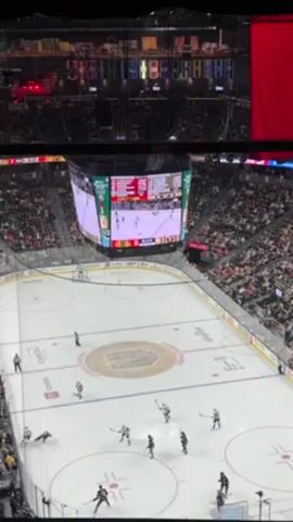 The video shows a wide shot of a hockey game in progress. The ice rink is brightly lit and surrounded by a large crowd of spectators. A large digital scoreboard hangs above the center of the rink, displaying the game score and other information. Several players in black and white uniforms are skating on the ice, along with a referee. The game is in motion, with players passing the puck.