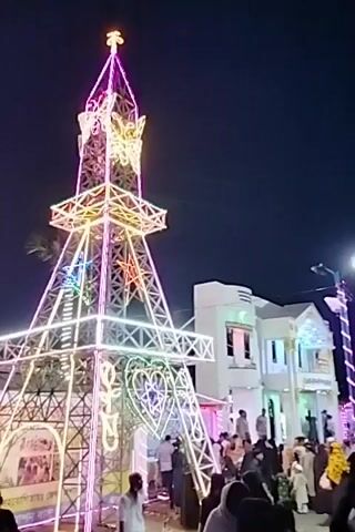 The video captures a lively night scene in Charfasson, Bangladesh, centered around two large, illuminated decorative structures. In the foreground, a prominent replica of the Eiffel Tower is brightly lit with a multitude of colorful LED lights, including pink, yellow, green, blue, and white. A large heart shape, also formed by lights, adorns its base, while stars are visible on its upper sections. Behind this structure, another tall, slender tower, similarly covered in colorful lights, stands out against the dark night sky.

A crowd of people is gathered around the base of the Eiffel Tower-like structure, walking and observing the festive display. The scene suggests a fair, carnival, or public celebration. Palm trees are partially visible to the left. In the background, a building with a red roof and white walls can be seen. On the building's facade, text in a local script is partially visible, appearing to say "স্বাগতম মিলাদ মাহফিল ও র... প্রাঙ্গণে..." which translates to "Welcome Milad Mahfil and R... Ground/Arena...". The overall mood is festive and vibrant, illuminated by the elaborate light displays.