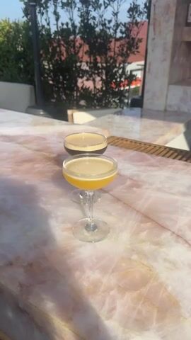 Two cocktail glasses, one with a yellow liquid and the other with a dark liquid with a foamy top, sit on a pink marble countertop outdoors. The sun is shining and reflecting off the surface. In the background is a blurred view of greenery and a building.