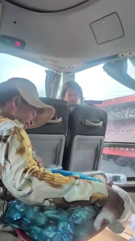 The video shows an interior view of a bus, likely in transit, as evidenced by the movement and passing scenery visible through the windows. The primary focus is on two individuals seated in the foreground and mid-ground. In the foreground, a person wearing a straw hat and a patterned shirt is visible from the side. They appear to be looking down at something in their lap, which is covered by a blue plastic bag. Their left hand is holding the edge of the bag. Behind this person, in the mid-ground, a young child is seated, looking towards the camera. The child has dark hair and is wearing a pink top. The background shows the interior of the bus, including the backs of seats, curtains, and the windows. The overall lighting suggests it is daytime.