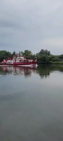 A red and white boat travels on a river under a cloudy sky. The boat has multiple decks and appears to be a fireboat, with red railings and what looks like a fire hose on top. Trees and vegetation line the riverbank in the background. The water is calm and reflects the boat and the sky. The boat is moving from left to right in the frame.