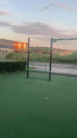 This video captures an outdoor fitness area with a calisthenics park as the main subject. The park features various bars and structures for exercise, all made of dark metal with bright green accents. The ground is covered in a soft, artificial green turf, likely for safety.

In the background, a road is visible, bordered by a strip of vegetation. Beyond the road, a modern, orange-colored building with large windows stands out, reflecting the soft light of the setting sun. Further in the distance, other low-rise buildings and a tall street lamp can be seen. A black metal fence runs along the right side of the frame, separating the fitness area from what appears to be a road or path.

The time of day is clearly sunset, indicated by the warm, diffused light and the soft hues in the sky, which are filled with scattered clouds tinged with pink and orange. The overall atmosphere is peaceful and serene, suggesting a quiet time for outdoor activity. There is no visible text in the video. The location is identified as Waalwijk, Netherlands. The scene is devoid of people, emphasizing the structures and the tranquil environment.