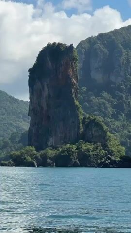A view from a boat shows a large, steep, tree-covered rock formation rising from the water. The rock is sheer on one side and has a more jagged, textured appearance on the other. Green vegetation covers the top and sides of the rock, as well as the larger mountainous landmass behind it. The water in the foreground is blue and choppy. The sky is partly cloudy with white clouds. The scene appears to be a tropical coastal landscape.
