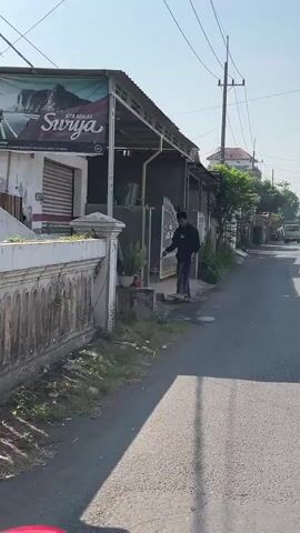 A man wearing a black long-sleeved shirt, black peci hat, and sarong is sweeping the ground in front of a building. He is holding a broom in his right hand and a dustpan in his left. There is a large potted plant to his left and a white wall with decorative elements behind him. The ground is paved and appears to be a sidewalk or pathway. The man is standing next to a concrete structure. The environment appears to be outdoors, possibly in a residential or commercial area, with a street visible in the background. The sun is shining, casting shadows.