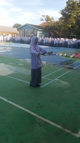 This media file captures an outdoor event at Rhieng Blang, Indonesia, likely a school gathering or ceremony. The scene is dominated by a green sports field with blue track lanes, indicating an athletic or event space. A bright, sunny sky suggests it's daytime, likely morning or afternoon.

The central subject is a young person, wearing a white hijab and a light-colored uniform with a red sash around their shoulders. This sash has the word "DIRI" printed on it in white letters. The individual is standing with their arms outstretched and raised, performing some kind of gesture or pose, possibly part of a dance or ritual.

In the background, a line of people, likely students, are standing on the track, all dressed in white uniforms and some wearing headscarves. They appear to be watching the central figure. Behind the students is a building, possibly a school or community hall, with a light blue facade and a red-tiled roof. Trees and vegetation are visible on the left side of the frame, adding to the outdoor setting.

The overall mood seems to be one of organized participation and potentially solemnity or celebration, given the formal attire and the structured arrangement of the people. The wet appearance of the track surface might suggest recent rain or a cleaning, though the bright sun indicates it's no longer raining. The image is oriented vertically, suggesting it was taken with a mobile phone.