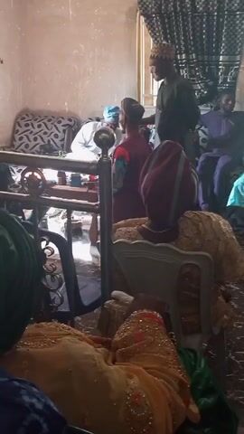 Okay, here's a detailed description of the video:

The video shows an indoor gathering of people, likely in a home. The setting appears to be a moderately furnished room. Several people are seated on sofas and chairs, while at least one person is standing and addressing the group. The individuals in the video are wearing traditional attire, including head coverings and colorful robes, suggesting a cultural or ceremonial event. A small wooden table with various objects on it is visible in the center of the room. The lighting in the room is moderate. The camera is mostly static, providing a view from the perspective of someone seated within the gathering. The focus is on the interaction between the standing person and the seated individuals.