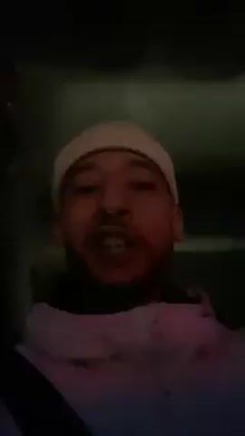 A man is recording a video of himself, illuminated by red light, while in what appears to be a car. He is wearing a white beanie and a light-colored jacket. The man's face is visible, and he appears to be smiling and speaking. A seatbelt is visible across his body. The background is dark and indistinct.