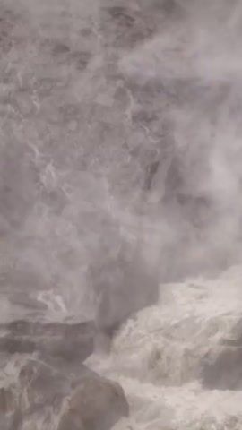 The video shows a powerful waterfall cascading over a rocky cliff face. The water is churning and creating a significant amount of mist and spray, obscuring some of the details of the rocks and the water's path. The overall impression is one of raw, natural power and the force of water in motion. The lighting is muted, contributing to the dramatic and somewhat mysterious atmosphere of the scene. The camera is positioned to capture the immense scale of the waterfall and its surrounding environment.