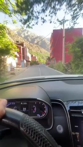 The video shows the driver's perspective of a car moving slowly down a narrow paved street in a village nestled in a mountainous area. The buildings on either side of the street are primarily two to four stories high and painted in shades of orange, peach, and red. In the distance, snow-capped mountains rise sharply against a clear blue sky. The driver's hands are visible on the steering wheel, which has a carbon fiber-like finish. The dashboard of the car is modern, with a digital display showing speed and other information. The scene is bathed in bright sunlight, creating a clear and vivid view of the village and surrounding landscape.