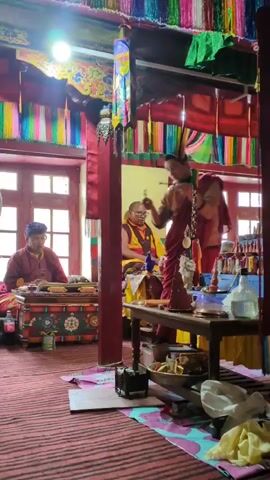 Two Buddhist monks are in a room with colorful prayer flags hanging from the ceiling. One monk on the left is seated and reading from a book. The other monk on the right is standing and holding a small object in his hands. There are several tables and religious artifacts in the room.