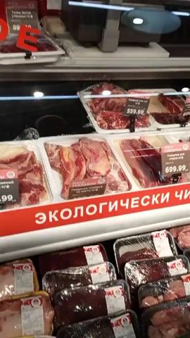 The video shows the inside of a grocery store. A refrigerated display case full of packaged meat products is visible, followed by a view of an aisle with shelves stocked with goods. Two people, a man and a woman, are walking down the aisle pushing a shopping cart. The camera pans up, showing the ceiling of the store with fluorescent lights. The camera is handheld and the footage is somewhat shaky.