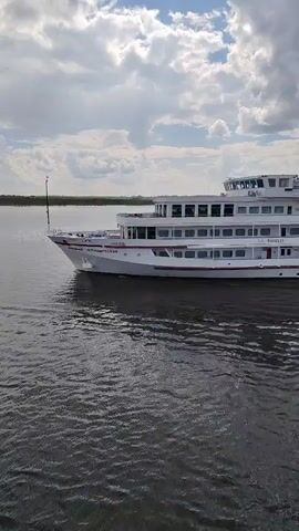 The video shows a white, multi-level river cruise ship moving on a body of water, presumably a river or a large lake. The ship, identified as "Владимир Маяковский" (Vladimir Mayakovsky), has multiple rows of windows and a raised navigation bridge on top. The water is dark and slightly rippled, suggesting a mild current or wind. The sky above is partly cloudy, with patches of blue sky visible between the clouds. The shoreline is visible in the background on the left side of the frame. The camera angle is slightly to the side of the ship, capturing its movement through the water.