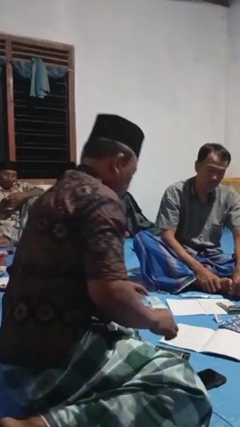 The video shows four individuals sitting on a blue tarp covering the floor of a room. Two men are in the foreground, one facing away from the camera and the other facing the camera but with his head down. Both are wearing sarongs. In the background, another man in a sarong and a black cap is visible, and a child is also in the background wearing a black garment and a cap. On the floor between the men in the foreground, there are papers, a calculator, a phone, a notebook, and a small container with what appear to be snacks or food. A small table with some items on it is visible against the wall in the background. The room appears to be dimly lit.