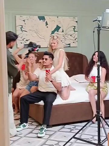 A group of people are in a room that looks like a bedroom, taking photos or filming a video. There is a man in the center wearing sunglasses, a striped shirt, and dark pants, holding a red cup. He is seated on a bed. Behind him are several women, some sitting on the bed and one kneeling behind him with her hands on his shoulders. Everyone appears to be holding a red cup. There is a lighting setup with a tripod and light facing the group. The room features a large bed with a brown headboard, a patterned floor, and a piece of art with floral designs on the wall behind the bed. The wall is a pale greenish color. A camera is visible on the left side, recording the scene. The view then pans to the right, showing more wall and part of a bed, but blurry.